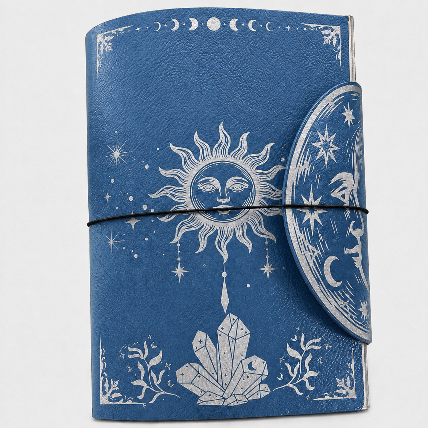 A blue journal with a wrap-around strap closure, decorated with white celestial and crystal illustrations including a sun face, moon phases, stars, and crystals, shown on a light gray background.